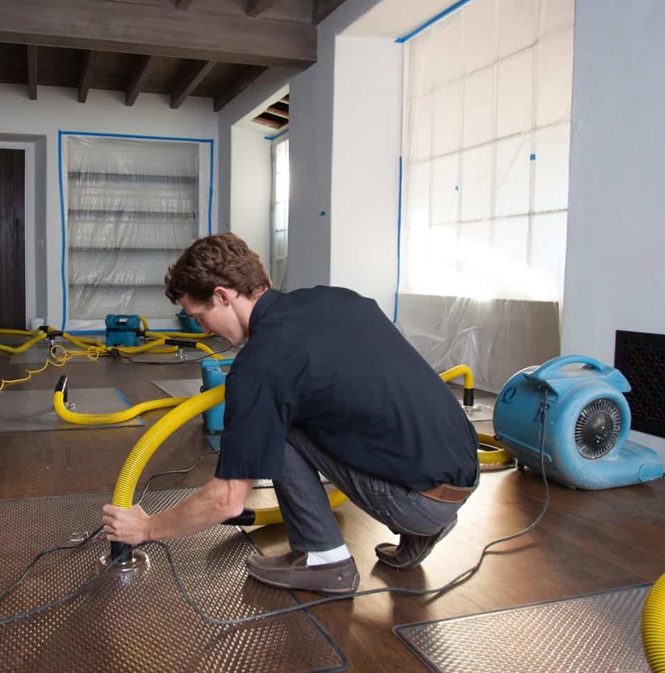 Prime Restoration technician running structural drying mats, hose, and air mover on hardwood after a water loss, with plastic containment taped to the windows